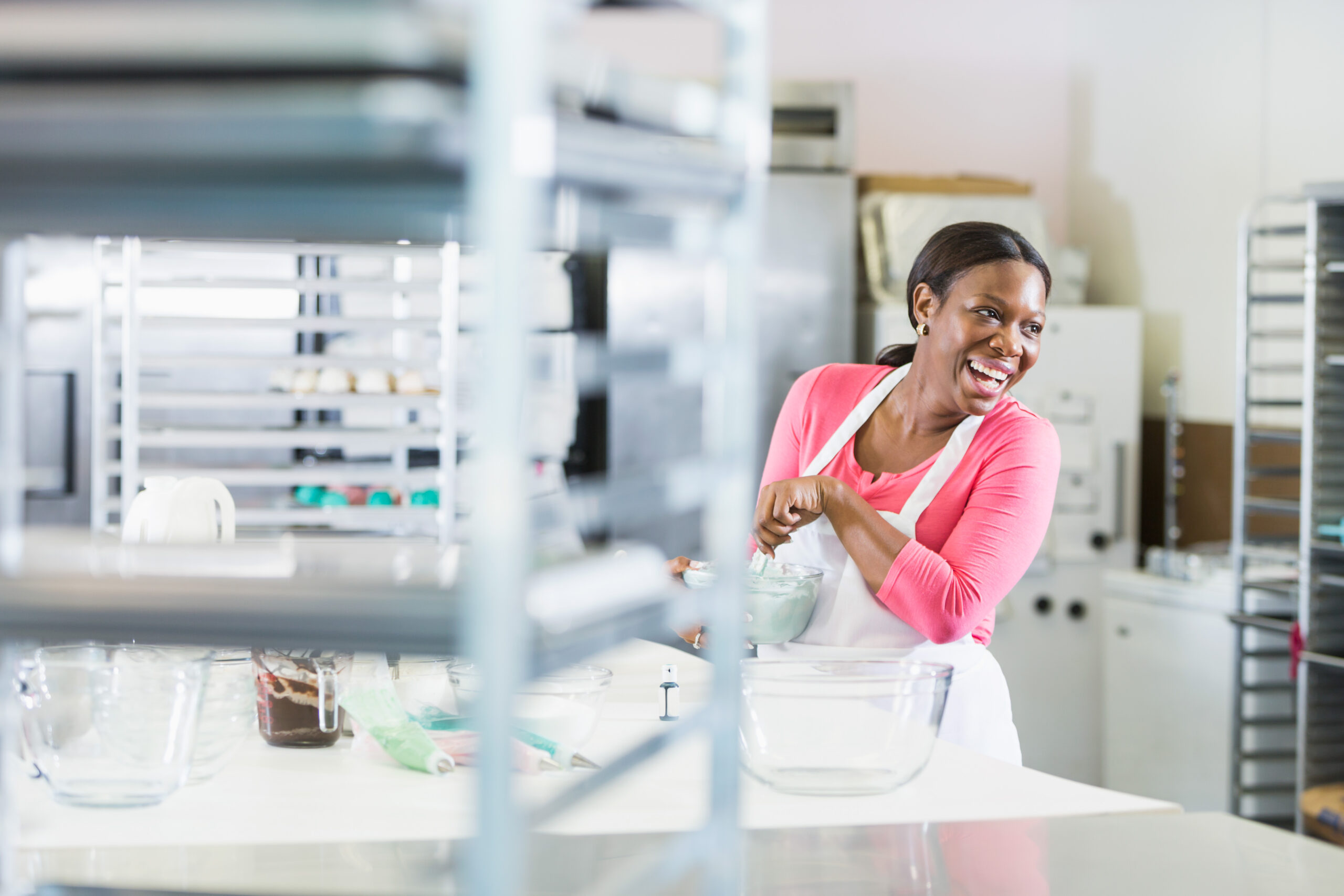 An African American woman wearing an apron, working in the commercial kitchen of a bakery. She is stirring a bowl, surrounded by baking racks and equipment. She is a successful minority small business owner.