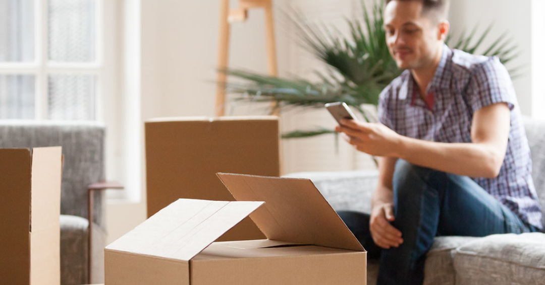 Man sitting on couch with boxes near him while he's looking on his phone for moving help.